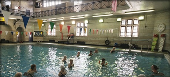 Aged photo of an indoor pool with multicolored flags hanging across the pool and a group of seniors in lines in the pool for a class