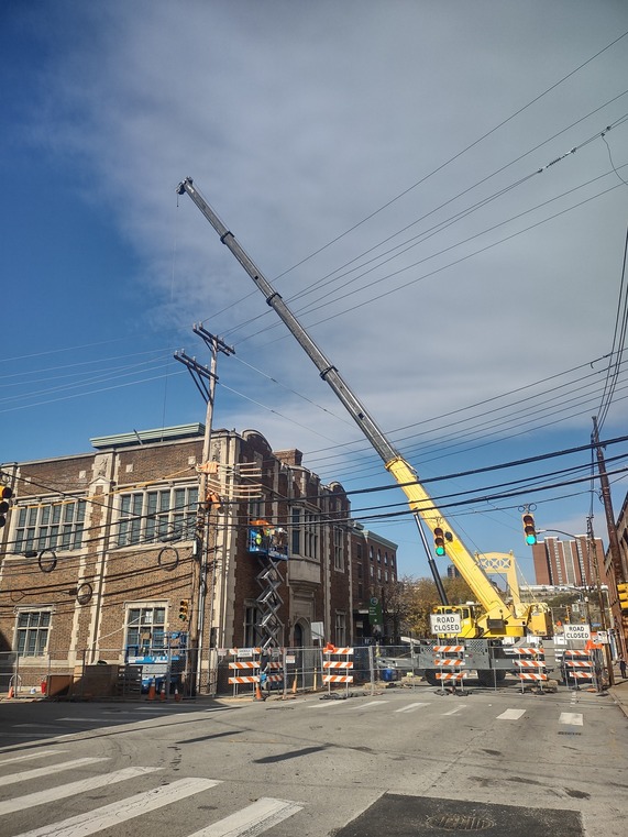 View from the street of road closed signs in front of a building with a large crane and construction workers in a scissor lift next to it