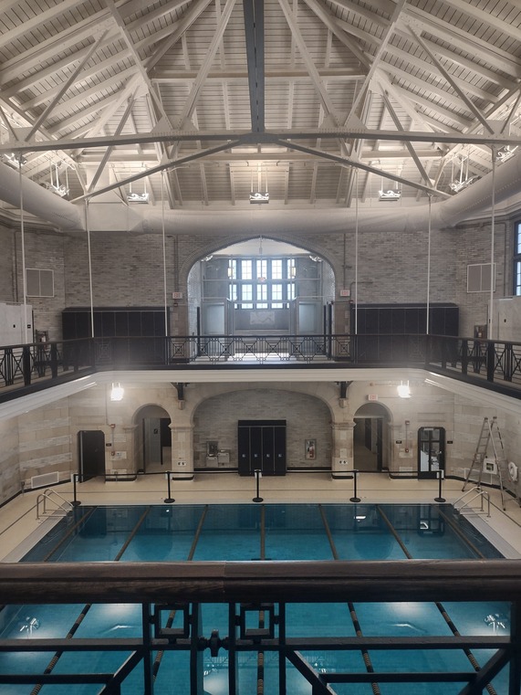 Interior of a pool building with high ceiling supported by white beams looking down on a pool with blue tile and walls and floors in white marble