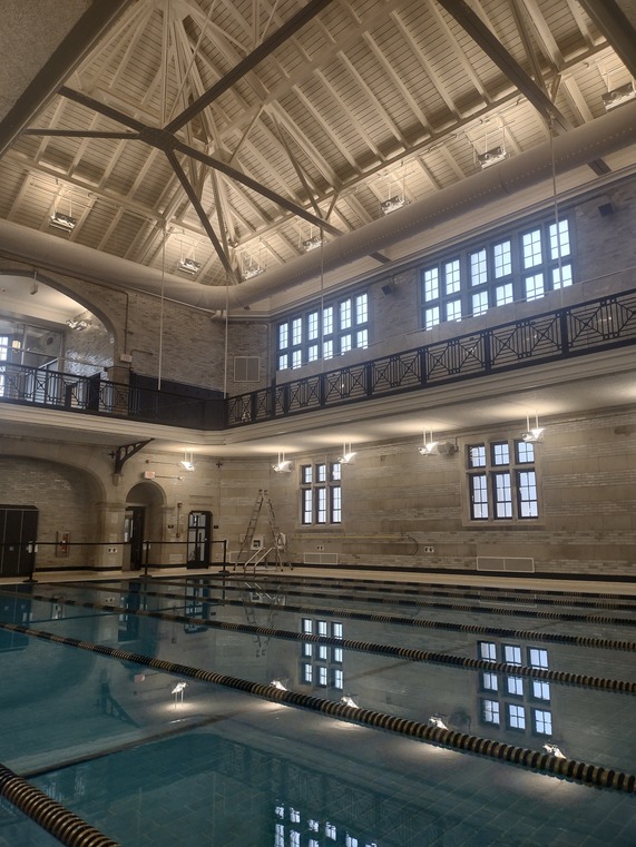 View from pool deck of blue indoor pool, deck has white and gray walls, black railings on the second floor and a white vaulted ceiling