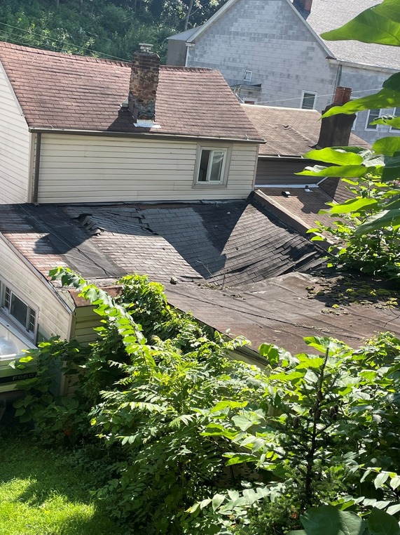 photo of the back of a two-story split level home with the back roof in the foreground caving in surrounded by weeds and overgrowth