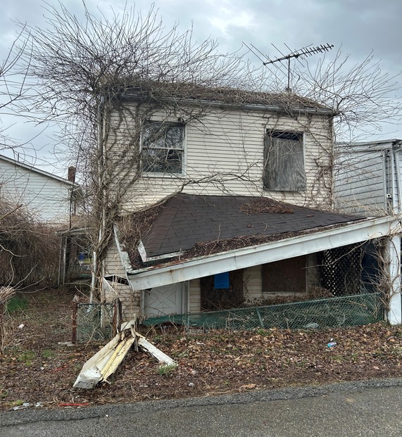 Photo of a two story house with trees growing along the wall and off the roof, the porch awning caved in and windows boarded up