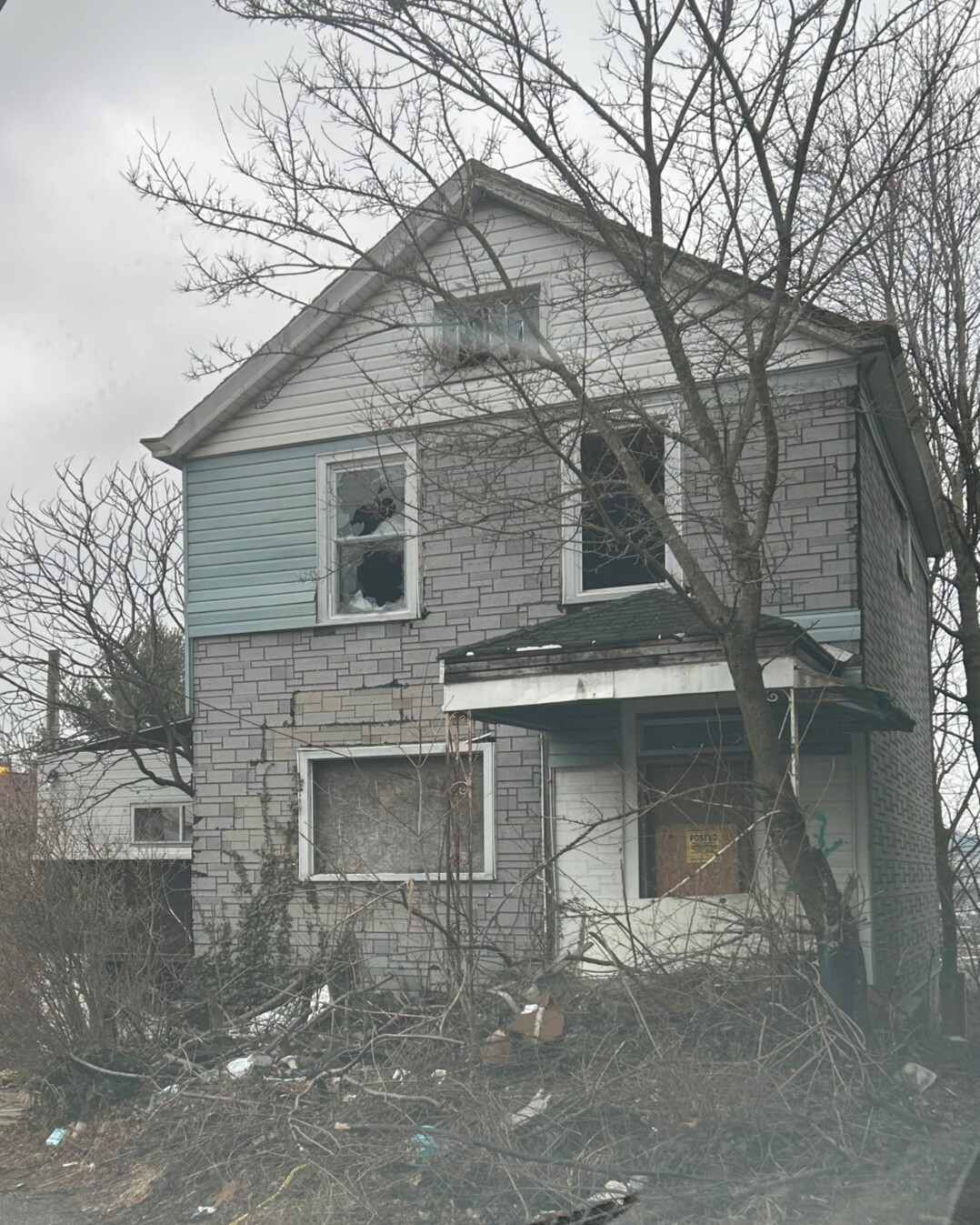 photo of a house with first floor window and door boarded, second floor windows broken and yard full of overgrowth and litter