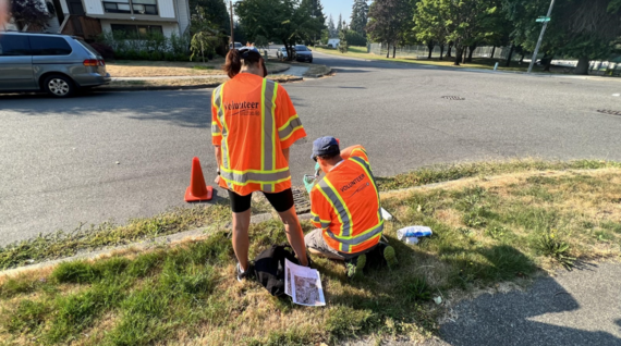 two volunteers in vests on a curb strip preparing a storm drain marking