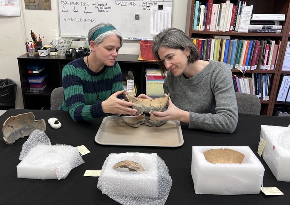 Drs. Beth Bollwerk and Lindsay Bloch examine a mended colonoware vessel at a table with bookshelves behind them.