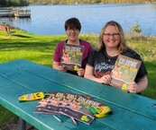 two people on a table with a lake in the background holding up a color book