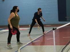 Two people wearing masks play pickleball in a gym. 