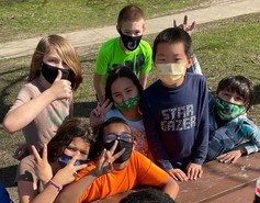Youth wearing masks and giving the thumbs up sign outdoors at a picnic table. 