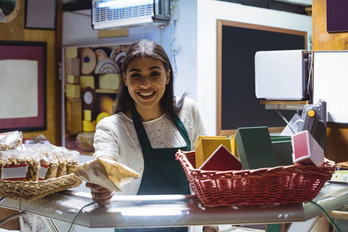 Smiling shop worker in an apron hands a paper-wrapped item to a customer over the counter, surrounded by snacks and colorful packaging