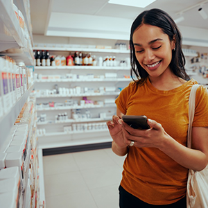 young woman smiling while using smart phone standing against shelf in pharmacy