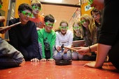 Wide shot of students observing experiment with goggles as teacher demonstrates