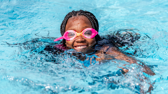 Black girl wearing pink goggles smiling for the camera while swimming.