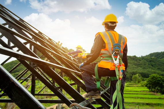 A construction worker stands on the framed roof of a house wearing a fall protection system.