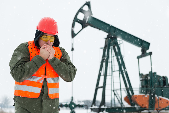 A man blows on his hands to warm them as he works in protective gear outside on a snow-covered refinery.