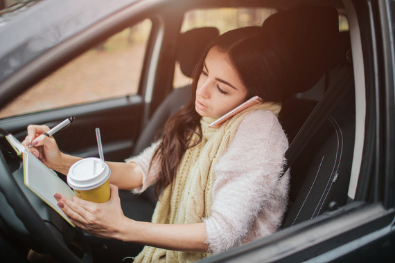Woman behind the wheel of the car eating, holding phone, and writing.
