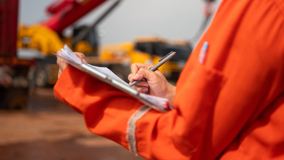 A man in orange coveralls writes on a clipboard as he stands in front of cranes.