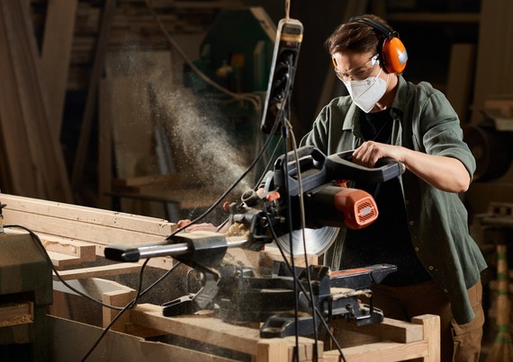 A man wears a dust mask and hearing protection as he uses a circle saw on a wood plank.