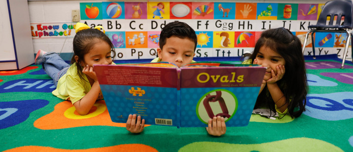 Three students on carpeted classroom floor, reading a book titled Ovals