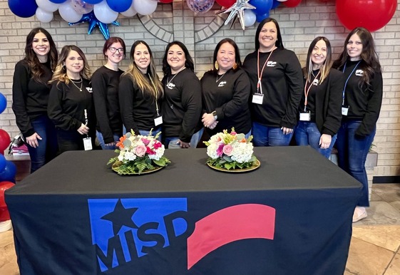 Ten people in black shirts stand behind a decorated table with the MISD logo and floral arrangements, in front of a patriotic balloon display.