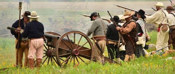 Group of historical reenactors in 1830s clothing, armed with rifles and gathered near a cannon in a field