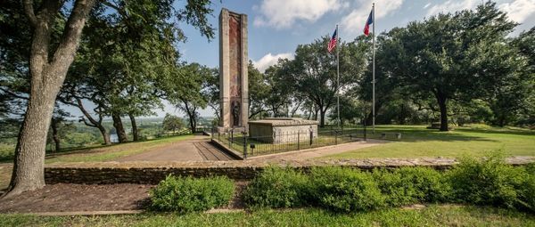 Tall Art Deco-style monument behind a large stone tomb at the top of a grassy hill, with the U.S. and Texas flags flying beside it