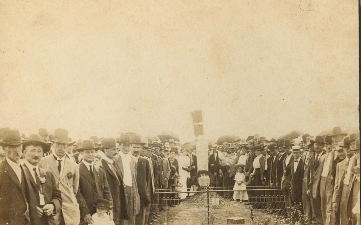A crowd stands around the grave marker where a pile of freshly turned dirt can be seen.