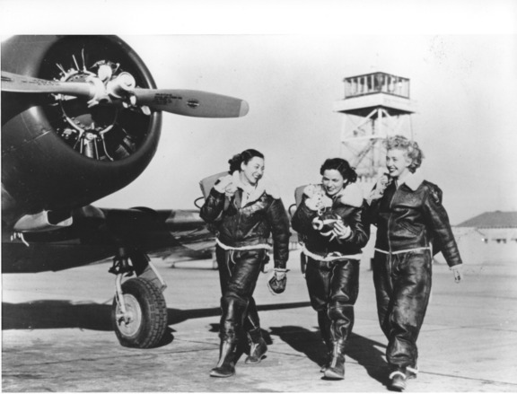 Three women in 1940s airforce attire laughing together and walking front of a plane.
