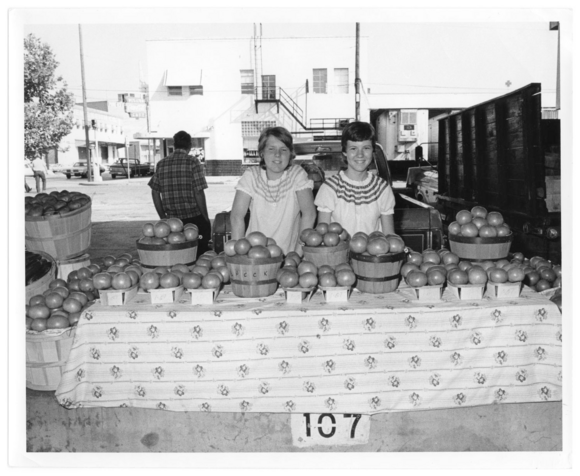 two girls displaying tomatoes at a farmers market table