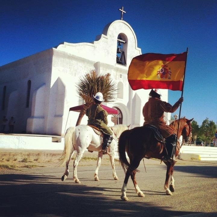 Two men on horseback. One is carrying the Spanish flag.