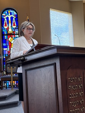 A woman wearing glasses is standing on the bimah at a synagogue presenting