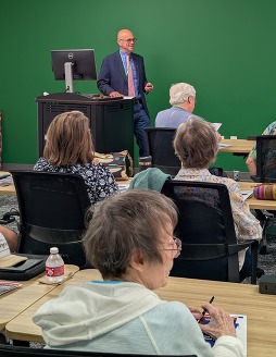 A man at a podium wearing glasses is presenting to a group of adults seated at tables. 