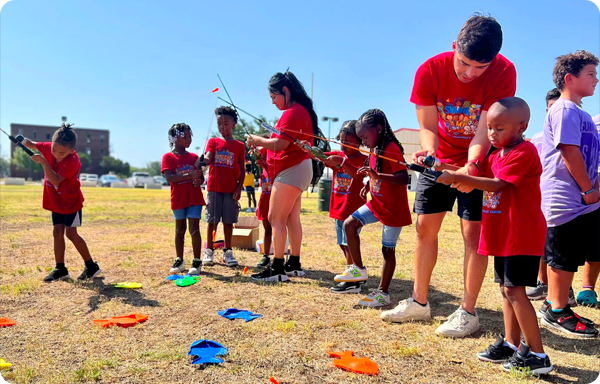 Two summer camp counselors helping kids with fishing rods in an open field.