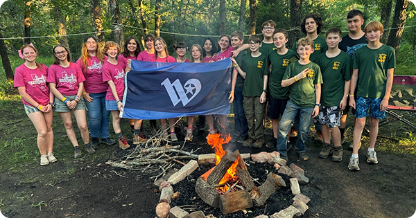 Group of kids behind a campfire in the woods holding the City of Waco flag