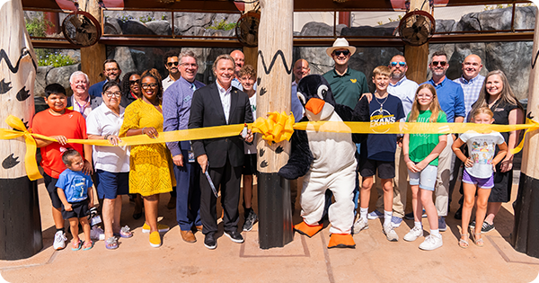 Group of men, women, and kids join the penguin mascot to cut ceremonial yellow ribbon in front of penguin exhibit