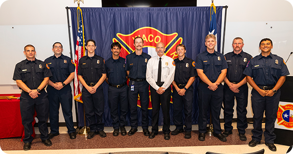 Nine fire recruits standing in a row with Fire Chief Scott Walker in front of a Waco Fire Department banner