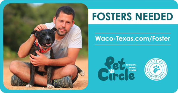 A man sitting cross-legged on a dirt road holding a happy black dog.