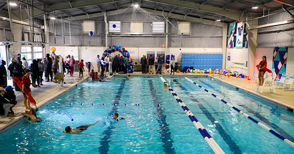 Kids swimming in pool. Lifeguards, people, and beachballs surround.