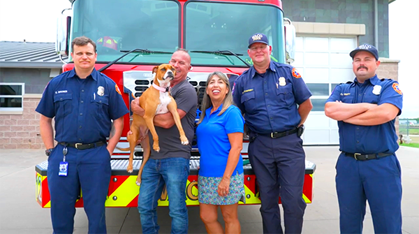 A happy couple holding their dog with three fire fighters