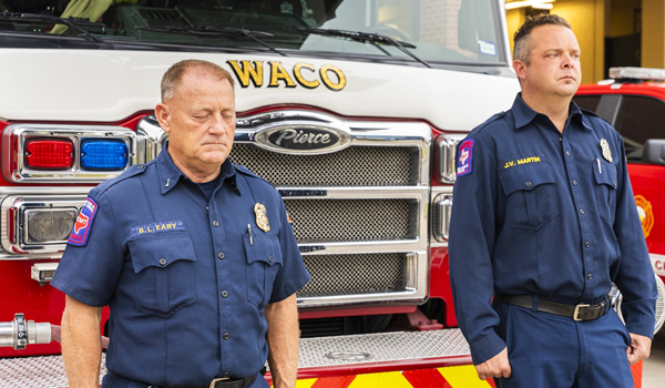Two firefighters standing somberly in front of a fire engine
