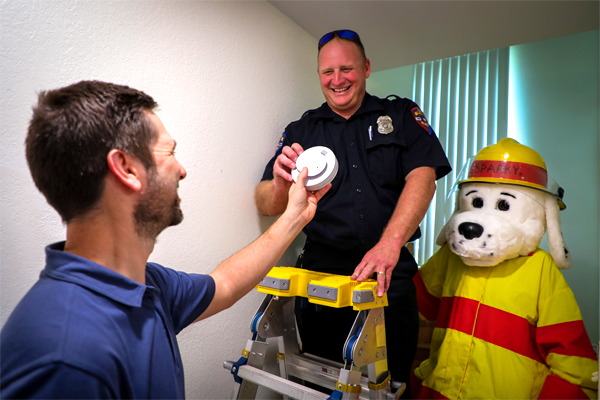 A man handing a fireman and Sparky the fire dog a smoke alarm