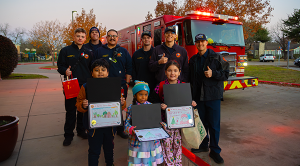 Firefighters standing with three kids holding their hand drawn Christmas cards