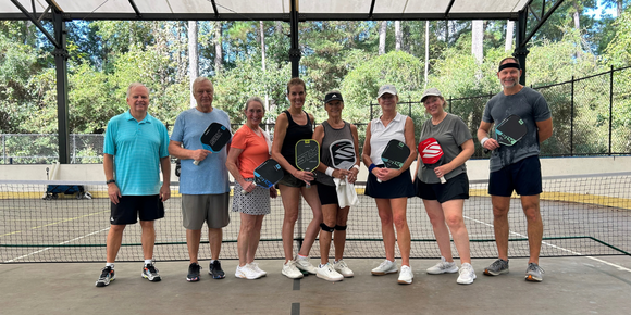 A group of people holding pickleball paddles dress in athletic gear standing on a covered pickleball court.