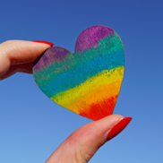 Rainbow coloured heart being held in a hand against a blue sky
