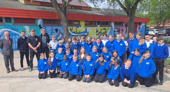 A group of children in blue jumpers and adults in front of a colurful yellow and blue mural