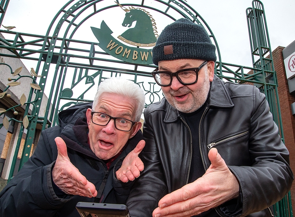 Two men in glasses looking at a phone in front of a sign saying Wombwell 