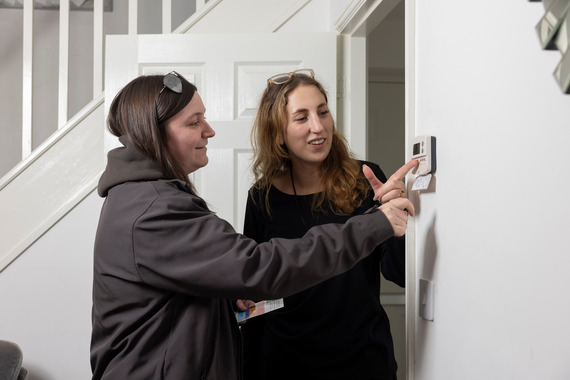 One woman is showing another woman how to use a thermostat on the wall.