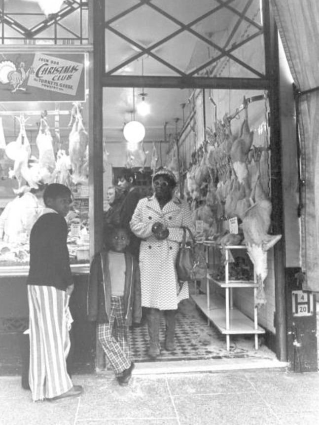 Photo showing 2 young black boys at doorway of butcher shop. Black woman stood inside shop. Poultry for sale hanging from hooks.