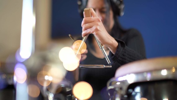 Artistic colour photo showing a musician about to strike a triangle (instrument) in a music studio. They're wearing headphones.