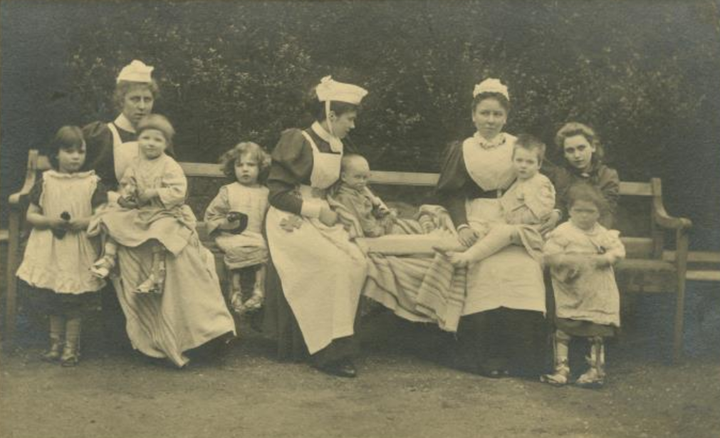 Black & white photo of nurses & children sat outside on a bench. The nurses are in uniform & the children appear to have physical disabilities.