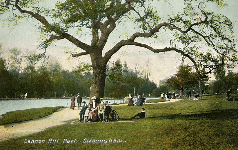 Colourised photo-postcard showing a footpath along a lake, a tree in the foreground and people walking & sat on the grass & benches.